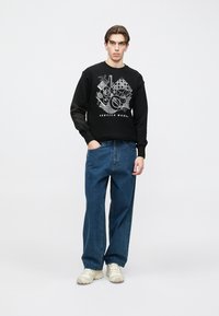 Μαύρο φούτερ με λευκό γραφικό σχέδιο, που αναγράφει το κείμενο "SERVICE WORK." Συνδυασμένο με μπλε φαρδιά τζιν και ανοιχτόχρωμα αθλητικά παπούτσια.