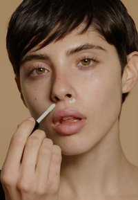 Påføring av ansiktspleieprodukt. Viser en lysfarget krem som påføres med en applikator på nesen. Jevn hud, kort mørkt hår.