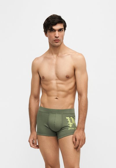 Jeune homme aux cheveux foncés portant un boxer vert olive avec un logo jaune, debout devant un fond clair uni.
