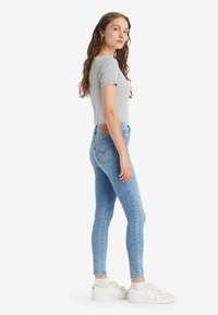 Νεαρή γυναίκα με μακριά σγουρά καστανά μαλλιά, φορώντας γκρι μπλούζα κοντομάνικη, ανοιχτό μπλε στενές τζιν, λευκές κάλτσες και λευκά αθλητικά παπούτσια, στέκεται στο πλάι.