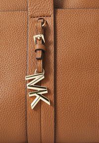 Borsa a tracolla in pelle marrone con una finitura texturizzata, caratterizzata da un charm con il logo 'MK' in tono oro attaccato da una tracolla coordinata.