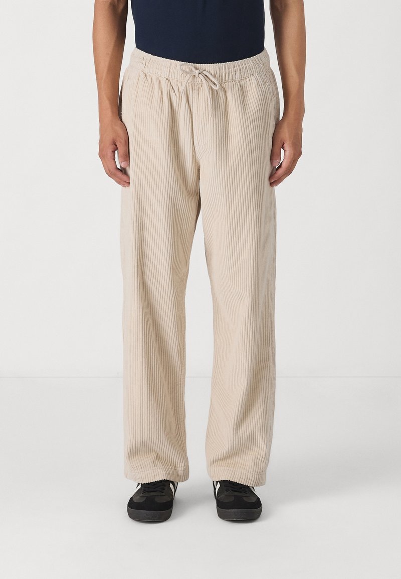 Beżowe spodnie sztruksowe o luźnym kroju, z pionowymi prążkami, elastyczną talią ze sznurkiem oraz kieszeniami po bokach.
