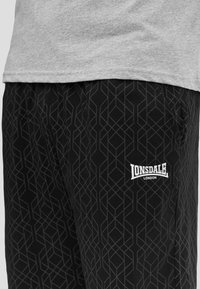 Zwarte broek met lichte geometrische lijnen en wit "Lonsdale London" logo op de linker dij, gecombineerd met een lichtgrijze zoom van het shirt.