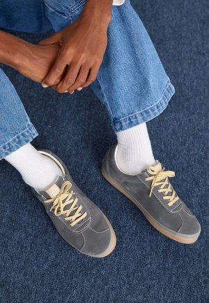 Grijze suède sneakers met beige veters en stitching. Rubberen zolen. Draag met witte sokken en denim broek. Handen rusten op de knieën.