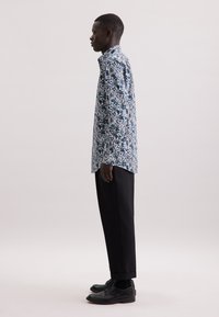Camicia a fantasia floreale blu e bianca, design con bottoni, abbinata a pantaloni e scarpe neri, mostrata in vista laterale.