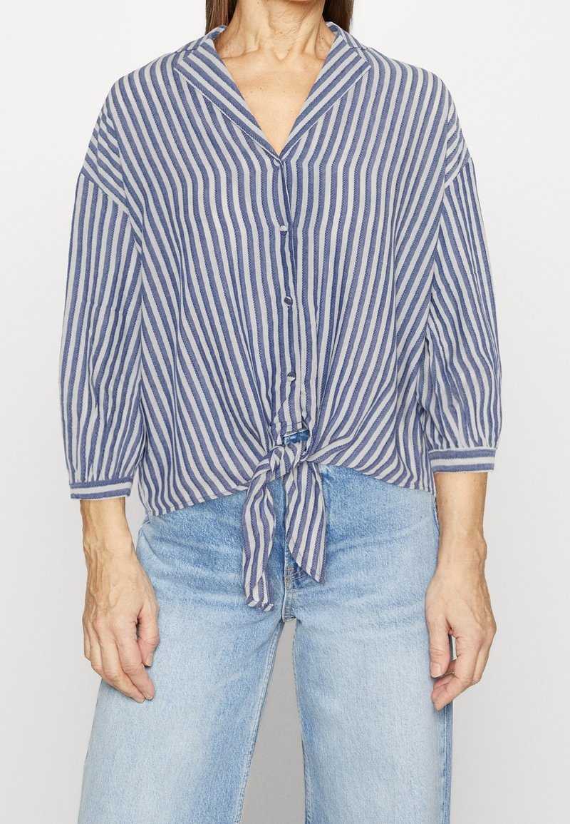 Femme portant une chemise à rayures verticales bleues et blanches, boutonnée, nouée à la taille, avec des manches trois-quarts et un jean bleu clair.