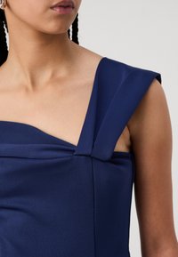Vestido azul marino con un escote estructurado y asimétrico; presenta un detalle de tela retorcida en el hombro y una textura suave y lisa.