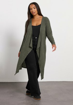 Offene Strickjacke aus olivgrünem Rippstoff, kombiniert mit einem schwarzen Oberteil und weiten schwarzen Hosen. Mit langen Ärmeln und einem fließenden Design.