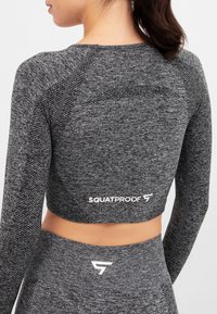 Grey long-sleeve athletic top with a textured knit pattern and fitted design. Features an elastic hem and "SQUATPROOF" logo at the back.