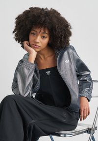 Metalno siva jakna na patentni zatvarač s elastičnim manžetnama, s malim bijelim Adidas logotipom. Ispod crni rebrasti komplet, sjedi na stolici.