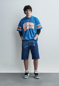 Ung mann står mot en grå vegg iført blå Billionaire Boys Club-drakt, denimbukser, hvite sokker og svarte og hvite joggesko.