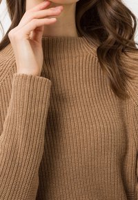 Pull en maille marron avec texture côtelée, manches larges et col haut ; le tissu semble doux et chaud.