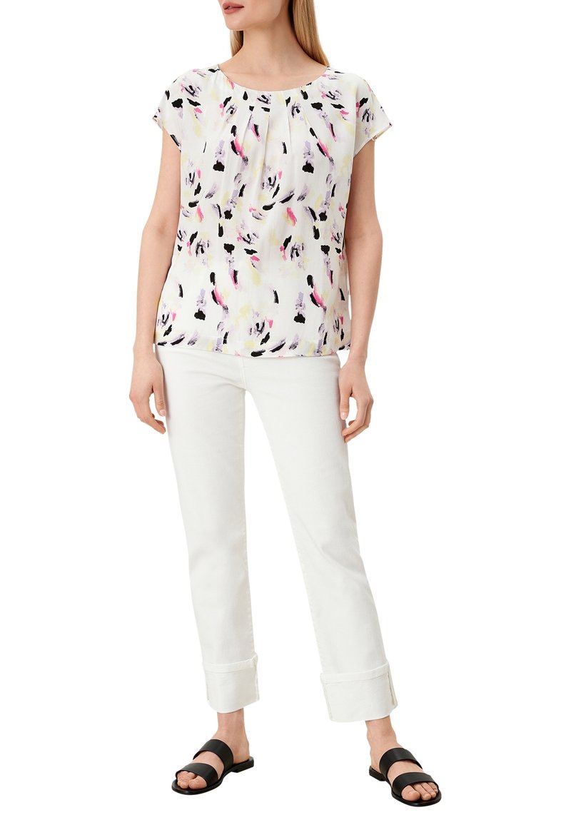 Femme portant un chemisier blanc avec des motifs en coups de pinceau noirs, roses et jaunes, un pantalon blanc avec revers et des sandales noires à bout ouvert.