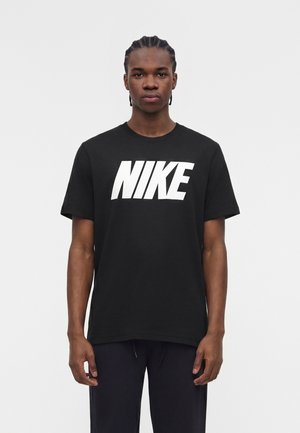 Junger Mann mit geflochtenem Haar, der ein schwarzes Nike-T-Shirt mit großem weißem Logo trägt und vor einem schlichten hellen Hintergrund steht.