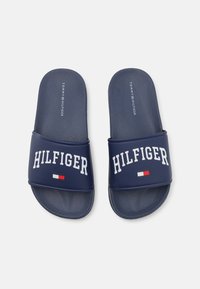 Chanclas de color azul marino con una superficie texturizada en la plantilla. Cuentan con una correa ancha con "HILFIGER" en blanco, adornada con una pequeña bandera italiana.