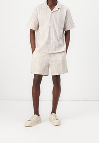 Camisa de manga corta de color beige claro a rayas, combinada con pantalones cortos a juego. La textura suave de la tela y las delgadas rayas contrastantes mejoran el diseño.