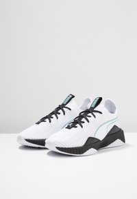 Scarpe sportive bianche con una tomaia testurizzata, lacci neri e una suola curva nera. Presentano accenti di blu chiaro e un logo prominente sulla linguetta.