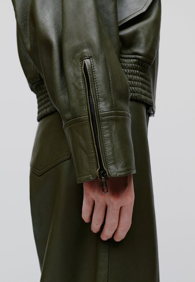 Prim-plan al unei mâini lângă fermoarul de la încheietura mânecii unei jachete de piele verde închis, cu detalii striate.