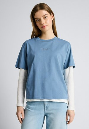 Młoda kobieta o długich włosach, ubrana w warstwowe niebieskie koszulki z krótkim rękawem i białe z długim rękawem oraz jasnoniebieskie dżinsy, stojąca na tle szarego tła.