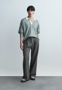 Fiatal férfi zsebre tett kézzel, világoszöld, texturált pulóvert, barna bőszárú nadrágot és kényelmes cipőt viselve.