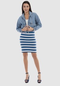 Jachetă albastră scurtă cu buzunare cu fermoar, top alb fără mâneci și fustă tip creion cu model zigzag în nuanțe de albastru și alb. Sandale negre cu toc înalt.