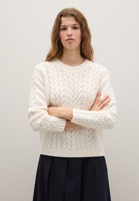 Cream knitted sweater with a cable pattern, round neckline, and long sleeves. Worn with a dark skirt, set against a light background.