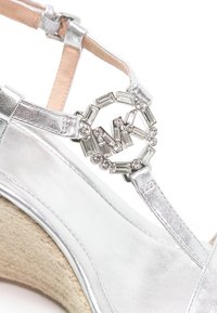 Sandale à talon compensé argentée avec bride de cheville et boucle circulaire ornée de strass sur fond blanc.