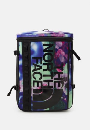 Sac à dos carré avec imprimé floral abstrait multicolore, sangles latérales noires, et grand logo vertical "The North Face" à l'avant.