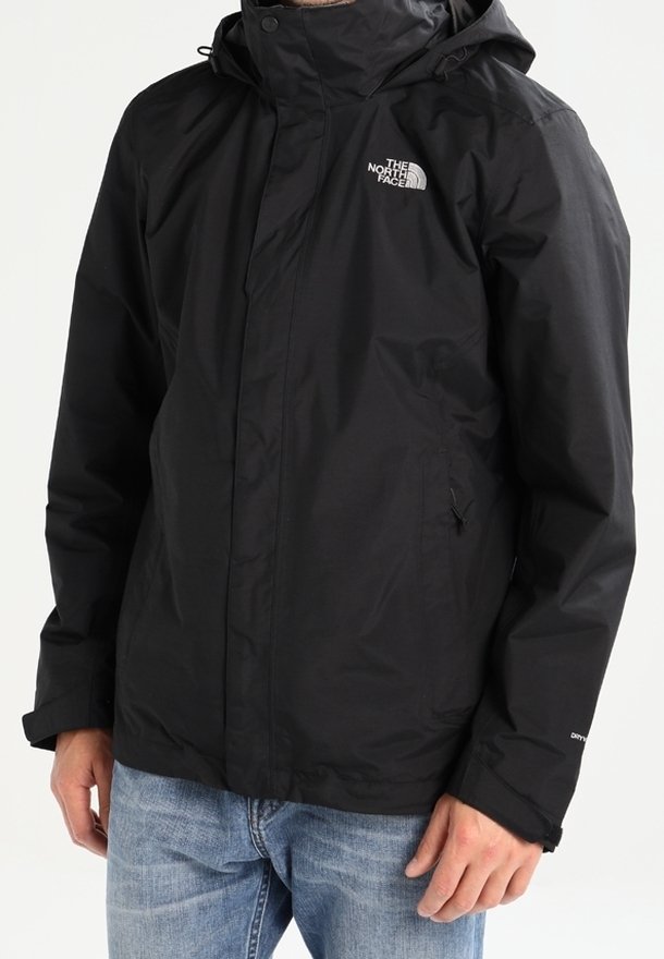 Homme portant une veste imperméable noire North Face avec capuche et poches zippées, associée à un jean bleu.