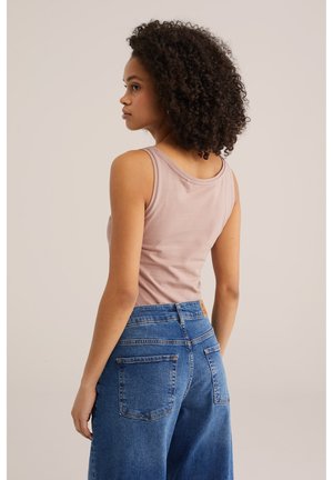 Kortsude juustega naine, kes kannab heleroosat varrukateta topi ja kõrge vöökohaga siniseid teksapükse, seisab ja vaatab küljele ühevärvilise tausta ees.