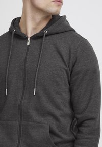 Donkergrijze rits-hoodie van zachte stof, met een voorzak, verstelbare trekkoorden en zilveren metalen details.