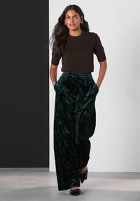 Femeie cu păr lung și închis la culoare, îmbrăcată cu un top negru cu mâneci scurte, pantaloni largi din catifea de culoare verde închis cu modele și pantofi ascuțiți de culoare albastră marin cu fundițe.