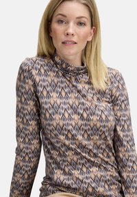 Blonde Frau, die ein langärmliges Oberteil mit hohem Kragen in einem braunen, grauen und beigen Rautenmuster vor einem weißen Hintergrund trägt.