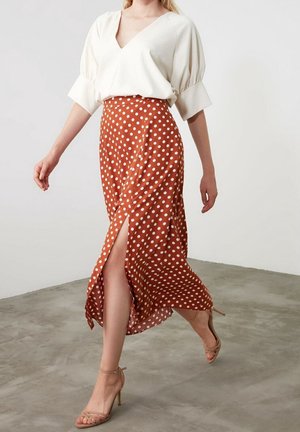 Femme marchant en blouse blanche à col en V et jupe taille haute orange rouille avec des pois blancs et une fente frontale, portant des talons beige à brides.