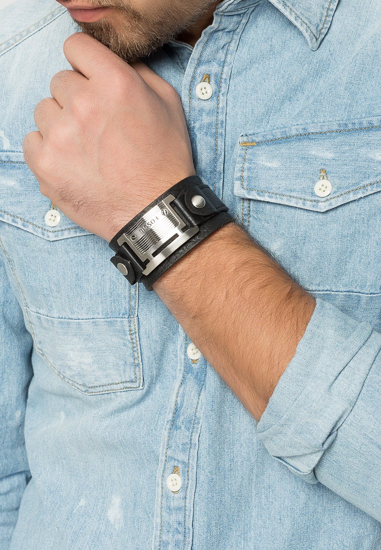 Bracelet en cuir noir avec accent en métal argenté arborant le nom de la marque. Porté au poignet, associé à une chemise en denim bleu clair.