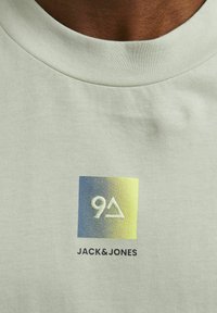 Camiseta verde claro con cuello redondo. Presenta un logo cuadrado impreso con colores en degradado (azul a amarillo) y el texto blanco "JACK & JONES".