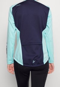Chaqueta atlética ajustada que presenta una parte trasera azul marino y mangas azul claro, hecha de tela suave con un logo en la manga izquierda.