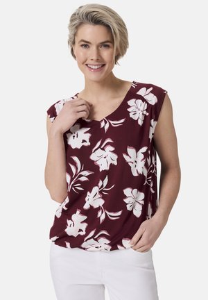 Bordeauxfarbene Bluse mit kurzen Ärmeln, die weiße Blumenmuster zeigt, aus einem weichen Stoff gefertigt ist und ein V-Ausschnitt-Design besitzt. Kombiniert mit weißen Hosen.