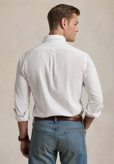 Λευκό πουκάμισο με μακριά μανίκια, γιακάς με κουμπιά, υφή υφάσματος, μανίκια γυρισμένα, σε συνδυασμό με μπλε τζιν και καφέ δερμάτινη ζώνη.