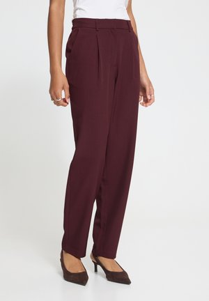 Personne portant un pantalon bordeaux plissé taille haute, un haut blanc et des escarpins bruns pointus, debout devant un fond blanc uni.