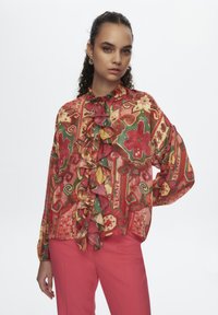 Mehrfarbige Bluse mit Rüschendetail an der Vorderseite, die florale und geometrische Muster in Rot, Grün und Gelb auf einem leichten Stoff zeigt.