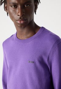 Violet spalvos megztinis su apykakle, pagamintas iš minkštos medžiagos, su briaunotais akcentais ir mažu juodu "BOSS" logotipu ant krūtinės.