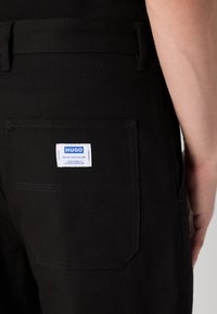 Black fabric trousers feature a back pocket with a white label reading "HUGO." The pocket has a rectangular shape and is stitched securely.
