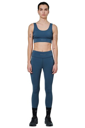 Kvinde stående iført blå sports-bh og leggings med sorte sokker og sko, tatoveringer synlige på arme, neutralt udtryk, hvid baggrund.
