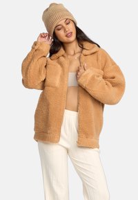 Giacca in felpa fluffy color nocciola con colletto, due tasche frontali e maniche lunghe. Indossata sopra una maglietta beige con pantaloni a strisce bianche e verticali.