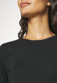 Czarny top z okrągłym dekoltem wykonany z miękkiego materiału. Zbliżenie ukazuje gładką fakturę, brak widocznych wzorów lub akcentów, oraz luźny krój wokół szyi.