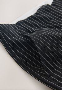 Schwarzer Stoff mit dünnen weißen vertikalen Nadelstreifen, zeigt gefaltete Kanten und eine kleine Schlitz-Detail.