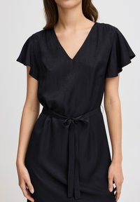 Schwarzes Kleid mit kurzen, fließenden Ärmeln, V-Ausschnitt und einem gebundenen Taillengürtel. Hergestellt aus glattem Stoff mit einem subtilen Glanz. Verfügt über seitliche Taschen.