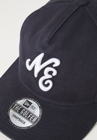 Tumesinine snapback müts, mis on valmistatud tekstureeritud kangast, valgete broderitud tähtedega "NE" ja ümmarguse sildiga, millel on kirjas "THE GOLFER."