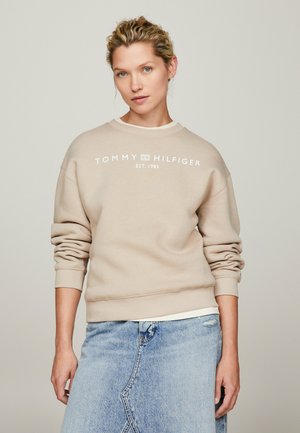 Kobieta o jasnych kręconych włosach nosi beżową bluzę Tommy Hilfiger i dżinsową spódnicę, stoi na tle jednolitego neutralnego tła.